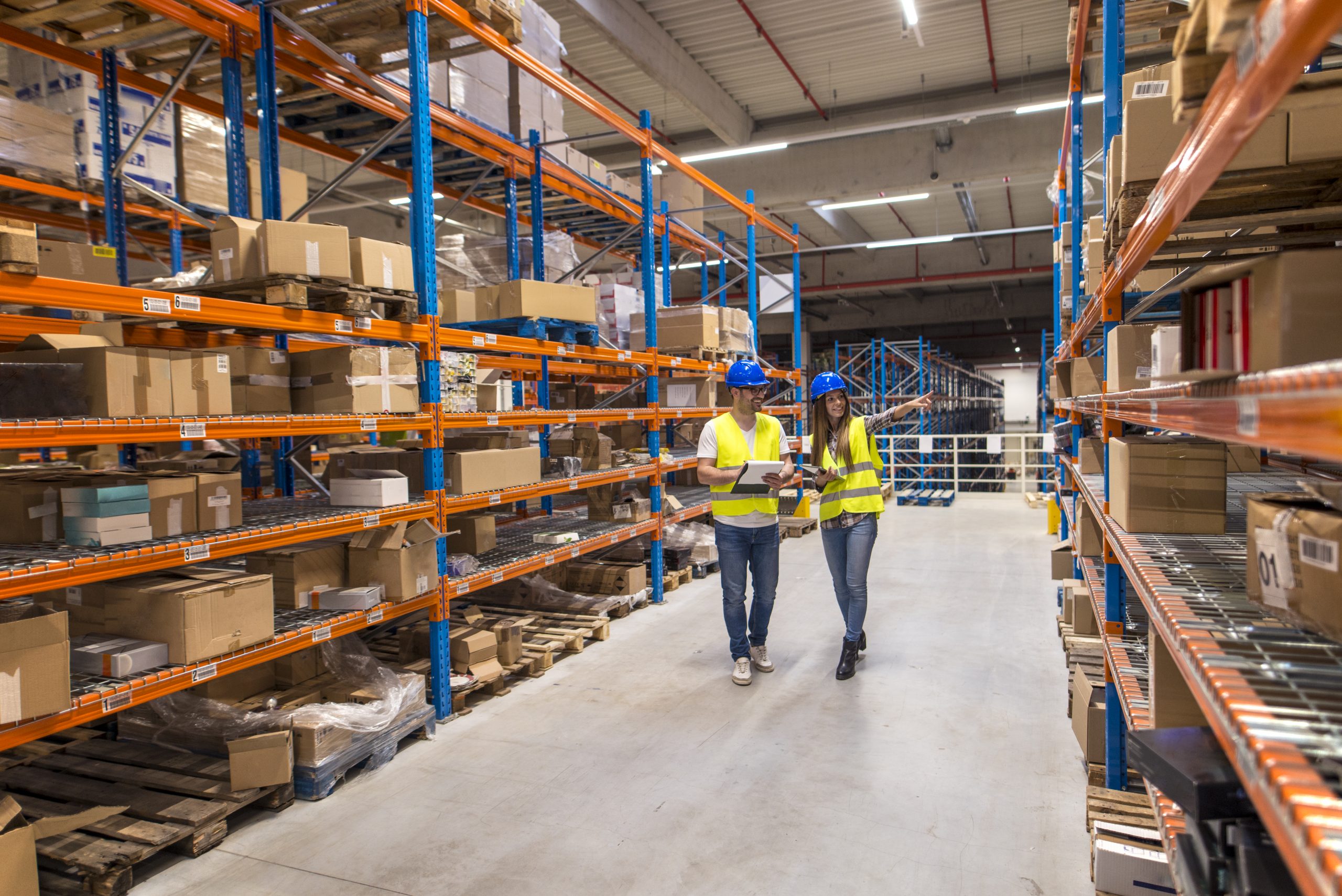 Two caucasian warehouse workers walking in distribution storage area discussing about logistics and organization.